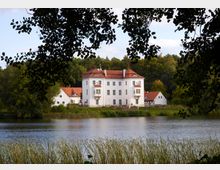 Historisches weißes Herrenhaus mit rotem Ziegeldach, umgeben von kleineren Nebengebäuden, steht am Ufer eines Sees. Im Hintergrund ist ein dichter Wald zu sehen, und Gräser säumen das Ufer im Vordergrund.