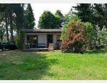 Kleines einstöckiges Haus aus Holz mit großen Fenstern, umgeben von einem Garten mit Rasen, Büschen und hohen Bäumen; links ist ein geparktes Auto sichtbar.