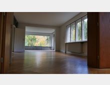 Großer, leerer Raum mit Holzboden, einer Tür auf der linken Seite und einer Fensterfront rechts, die Blick auf Bäume und herbstliche Landschaft bietet. Die Fenster lassen viel Tageslicht ein, unter den Fenstern befinden sich Heizkörper.