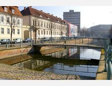 Historischer Straßenzug mit gepflastertem Gehweg, Blick auf einen Kanal mit einer kleinen Metallbrücke im Vordergrund. Im Hintergrund stehen Gebäude mit heller Fassade und roten Ziegeldächern sowie ein moderneres Hochhaus.