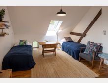 Ein gemütlich eingerichtetes Dachzimmer mit zwei Einzelbetten, einem Holztisch, einem Stuhl und einer Stehlampe. Ein Dachfenster spendet Tageslicht, Holzbalken und eine helle Wandgestaltung prägen den Raum.