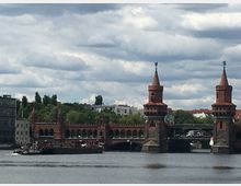 Backsteinbrücke mit zwei Türmen, bekannt als Oberbaumbrücke, über einem Fluss unter bewölktem Himmel; im Hintergrund Bäume und urbane Gebäudestrukturen, im Vordergrund ein Wasserfahrzeug.