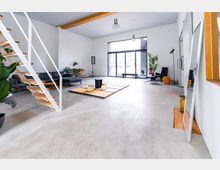 Moderner Wohnraum mit minimalistischem Design, sichtbar durch helle Wände, große Fensterfront und Betonboden. Eine offene Treppe mit Holzstufen, schwarze Ledermöbel, Pflanzen und ein flacher Holzpodest bilden die Hauptelemente der Einrichtung.