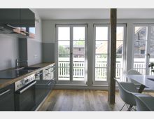 Moderne Küche mit einer Einbauküchenzeile in schwarzem Hochglanz, Holzarbeitsplatte und einem Fensterbereich mit drei großen Glas-Doppeltüren, die zu einem Balkon mit weißem Geländer führen. Der Raum hat Holzboden, eine freistehende Holzsäule und einen Essbereich mit weißen Stühlen.