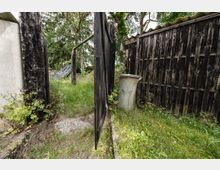 Schmaler, verwilderter Gartenweg zwischen dunklen, verwitterten Holzzäunen und einer halb geöffneten Holztür. Hohe Gräser, Bäume und ein grauer Metallmülleimer stehen neben einem Holzschuppen mit gewelltem Dach.