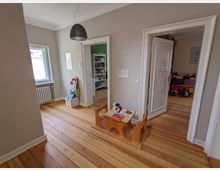 Heller Wohnungsflur mit breiten Holz-Dielen, grauen Wänden und schwarzer Pendelleuchte; links ein Fenster über einem Heizkörper. Zwei weiße Türrahmen führen in angrenzende Räume, im Vordergrund steht ein niedriger Kindertisch mit Bänken.