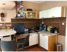 Kleine Küche mit heller Holz- und weißen Fronten, einer Arbeitsplatte mit kleinem Barbereich und zwei Barhockern. Zu sehen sind ein Herd mit Dunstabzugshaube, eine Mikrowelle, eine Kaffeemaschine, ein Wasserkocher sowie diverse Dekorationen wie eine Wanduhr und Notizen an der Dunstabzugshaube.