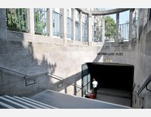 Eingang zur U-Bahn-Station Viktoria-Luise-Platz mit Treppen, Metallgeländern und einer gekrümmten Steinmauer; oben sind Balustraden und eine Beschilderung sichtbar, während eine Person mit einem roten Koffer den unteren Bereich betritt. Im Hintergrund sind Gebäude und Bäume zu erkennen.