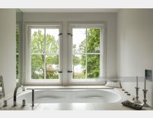 Helles Badezimmer mit einer eingebauten Badewanne vor einem großen, zweiflügeligen Fenster, das Blick auf grüne Bäume und einen See bietet. Auf der Fensterbank stehen Dekorationsgegenstände wie Kerzenhalter und kleine Steine.