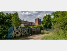 Backsteinbau mit Flachdach und Lüftungsanlagen, umgeben von Bäumen und einer Wiese, im Vordergrund eine mit Graffiti bemalte Backsteinmauer und ein kleiner Weg.
