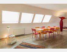 Ein heller Raum mit vier großen, schräg angeordneten Dachfenstern. In der Mitte steht ein Esstisch mit bunten Stühlen, daneben eine rote, hohe dekorative Lampe; der Boden ist aus Holz und ein gemusterter Teppich liegt im Vordergrund.