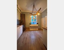 Geräumige Küche mit Holzboden, graugrünen Wänden und einem großen Fenster über einer Heizkörperverkleidung. An der Decke hängt eine rustikale Leuchte aus Treibholz mit mehreren Glühbirnen; links stehen weiße Unterschränke mit dunkler Arbeitsplatte und rechts eine Kücheninsel.