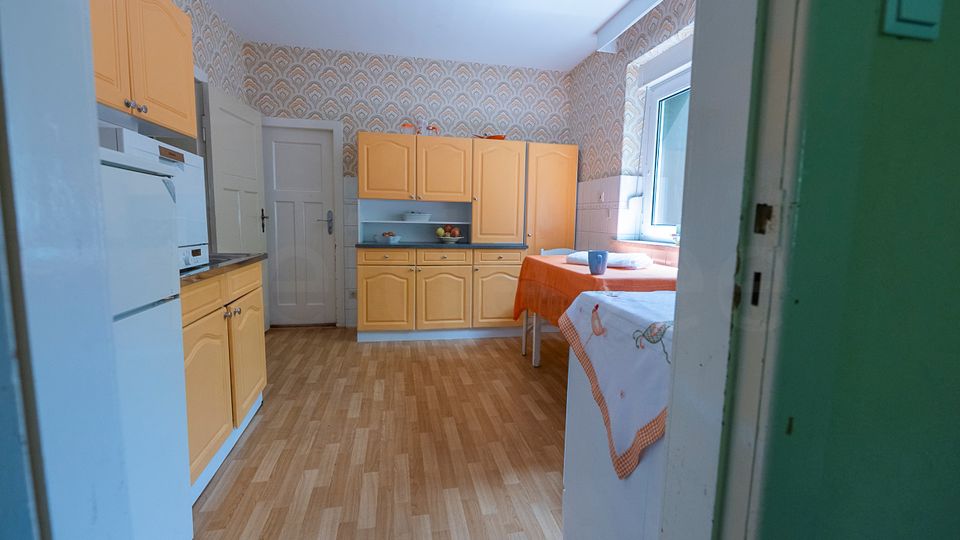 Blick in eine helle Wohnküche mit gelben Einbauküchenschränken, weißem Kühlschrank und Holzboden. Rechts steht ein Tisch mit orangefarbener Tischdecke neben einem großen Fenster; die Wände sind mit gemusterter Tapete und Fliesenspiegel versehen.