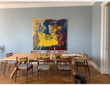 Esszimmer mit einem großen Holztisch und sechs Stühlen mit gemusterten Polstersitzen auf einem Parkettboden. An der blassblauen Wand hängt ein farbenfrohes abstraktes Gemälde in Gelb, Blau und Rot.
