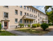 Mehrfamilienhaus in hellbeigem Putz mit drei Stockwerken und Satteldach, mehreren Fenstern und kleinen Balkonen; davor Grünflächen mit Büschen, ein gepflasterter Gehweg und ein Eingang mit roter Tür. Im Hintergrund Bäume und teils bewölkter Himmel.