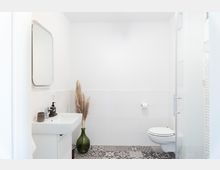 Minimalistisch eingerichtetes Badezimmer mit weißen Wänden und einer gemusterten Fliese auf dem Boden. Zu sehen sind ein wandhängendes Waschbecken mit Spiegel, eine Toilette, ein Glasvasen-Dekor mit Pampasgras sowie ein Glasausschnitt einer Duschwand.
