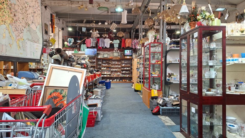 Innenansicht eines großen Secondhand- bzw. Trödelladens in einer Industriehalle mit hohen Decken, vielen hängenden Lampen und dicht gefüllten Regalen. Ein breiter Gang führt zwischen Vitrinen mit Glaswaren, Kisten, Einkaufswagen und ausgestellten Gegenständen bis zu einer Rückwand mit weiteren Regalen und aufgehängter Kleidung.