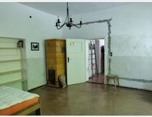 Ein einfacher Raum mit kahlen weißen Wänden, teilweise beschädigtem Putz und sichtbaren Reparaturspuren. Zu den Möbeln gehören ein Bett, ein gelber Kachelofen, ein Holzregal, ein Hocker und eine Deckenlampe mit schwarzem Metallrahmen; im Hintergrund ist eine Tür mit Blick in einen angrenzenden Raum erkennbar.