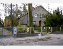 Ein zweistöckiges Einfamilienhaus mit grauer Fassade, grünen Fensterläden und Satteldach, umgeben von Bäumen und einem Garten. Im Vordergrund befindet sich ein Metallzaun mit einem Tor, das zur Straße führt.