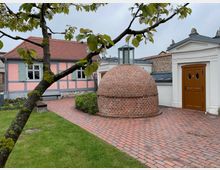 Ein kleiner Hof mit einem gepflasterten Weg, einem runden roten Ziegelbau mit Glaselement oben, und einer weißen Fassade mit brauner Tür. Im Hintergrund steht ein Fachwerkhaus mit rosa und grauen Wänden und grün gestrichenen Fensterläden, umgeben von einem gepflegten Garten.