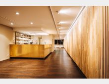 Ein moderner Innenraum mit einer langen Holzlamellenwand auf der rechten Seite, einem goldfarbenen Tresen auf der linken Seite und einer Deckenbeleuchtung mit eingebauten Spots. Der Boden besteht aus dunklem Holz, und im Hintergrund sind große Fenster sichtbar.