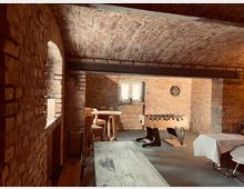 Kellerraum mit gewölbter Backstein-Decke, Wänden aus rotem Ziegelstein und Einrichtung im rustikalen Stil, darunter ein Tischkicker, ein Dartbrett, ein Holzhochtisch mit Hockern und ein Holztisch im Vordergrund. Ein kleines Fenster sorgt für Tageslicht.