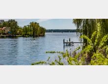 Ein See mit klarem Wasser, umgeben von Bäumen und Sträuchern; im Vordergrund ein kleiner Steg mit einem Vogel, im Hintergrund Segelboote auf dem Wasser und ein Haus am Ufer.