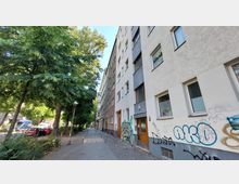 Straßenansicht mit mehrstöckigen Wohngebäuden auf der rechten Seite, deren Fassaden teilweise mit Graffiti versehen sind, sowie einem Gehweg und Bäumen auf der linken Straßenseite, die Schatten spenden. Im Hintergrund sind parkende Autos und eine beleuchtete Straßenlaterne sichtbar.