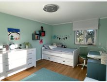 Ein Kinderzimmer mit mintgrünen Wänden, Holzfußboden und weißer Einrichtung. Zu sehen sind ein Bett mit Schubladen, ein Schreibtisch mit grünem Stuhl unter einem Fenster sowie eine Kommode mit Pflanzen und Dekoartikeln.