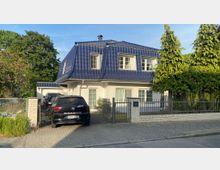 Zweistöckiges Einfamilienhaus mit weißer Fassade und dunkelblauem Walmdach, umgeben von einem Metallzaun und grüner Vegetation. Neben dem gepflasterten Eingangsweg sind Autos auf einem Parkplatz abgestellt.