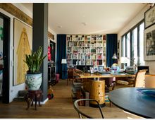 Wohnraum mit Bücherregal und Schreibtisch. Der Raum ist mit einem großen Regal voller Bücher, mehreren Möbelstücken im Vintage-Stil, großen Fenstern mit Tageslicht und Zimmerpflanzen dekoriert.