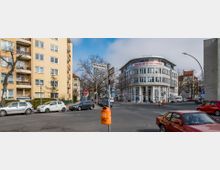 Straßenszene mit einem modernen, halbrunden Bürogebäude auf der rechten Seite, das mit "Hausarzt Germedica" beschriftet ist, sowie einem mehrstöckigen Wohnhaus mit gelber Fassade auf der linken Seite. Im Vordergrund befinden sich parkende Autos, ein Mülleimer und Straßenschilder, darunter ein Schild mit der Aufschrift "Ritterlandweg".