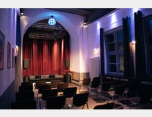 Ein kleiner Veranstaltungsraum mit einer Bühne im Hintergrund, flankiert von roten Vorhängen und Lautsprechern. Der Raum ist mit Reihen aus schwarzen Stühlen, hohen Fenstern mit Vorhängen und einer beleuchteten Discokugel über dem Bühnenbogen ausgestattet.