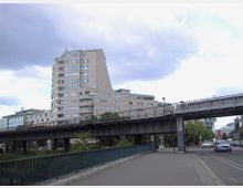 Mehrstöckiges Wohn- oder Bürogebäude mit abgerundeter Architektur im oberen Bereich, daneben eine erhöhte Brücken- oder Bahntrasse aus Stahl. Im Vordergrund sind eine Geh- und Radwegfläche sowie eine Straße mit Autos zu sehen.