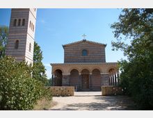 Eine Kirche im romanischen Stil mit einer Fassade aus hellem Ziegelstein und einem Rosettenfenster, flankiert von einem hohen Glockenturm. Der Eingangsbereich besitzt mehrere Rundbögen, und die Umgebung ist von Bäumen und Sträuchern geprägt.