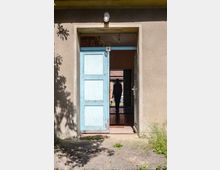 Blick auf eine geöffnete, hellblaue Holztür in einem Gebäude mit beige verputzter Fassade. Im Türdurchgang ist eine Person als Silhouette zu sehen, dahinter ein Innenraum mit Fliesenboden und ein weiteres Gebäude im Hintergrund.