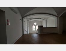 Ein heller Innenraum mit weiß verputzten Wänden, gewölbter Decke und einem Treppenbereich. Großzügige Fenster mit Rundbögen und Kreisformen lassen Tageslicht herein, daneben befinden sich Fluchtweg- und Toilettenschilder.