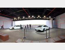 Großer moderner Ausstellungsraum mit zwei weißen Elektroautos, die auf einem beleuchteten Display präsentiert werden. Der Raum hat eine minimalistische Gestaltung mit roten LED-Linien an den Wänden und eine offene Glasfront mit Blick auf ein weitläufiges Gelände.