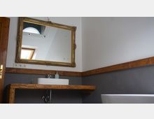 Badezimmer mit einem rechteckigen Waschtisch aus Holz, auf dem ein weißes Waschbecken steht, darüber ein großer, vergoldeter Spiegel. Im Hintergrund eine schräge Decke mit Oberlichtfenster und Wandverkleidung aus Holz und grauem Putz.