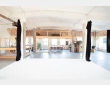 Großer, offen gestalteter Loft-Raum mit hohen Decken und sichtbaren Ziegelwänden. Im Hintergrund befinden sich eine hölzerne Empore, eine lange Tischgruppe, große Fenster und Pflanzen; schwarze Vorhänge rahmen den Bereich ein.