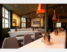 Innenraum eines modernen Restaurants mit langen, weiß gedeckten Tischen, gepolsterten Stühlen und großen Fenstern entlang der linken Wand. Über den Tischen hängen kupferfarbene Pendelleuchten; im Hintergrund sind Säulen, Wandkunst und weitere Tischreihen zu sehen.
