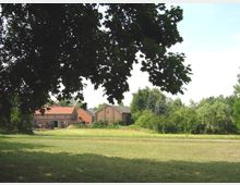 Eine ländliche Szene mit mehreren Ziegelgebäuden und roten Dächern, umgeben von Bäumen und Wiesen. Im Vordergrund ist eine große grüne Rasenfläche, teilweise im Schatten eines Baumes.