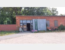 Ziegelsteingebäude mit einem Hofladen-Eingang, der von großen Glastüren und einem grünen Schild mit der Aufschrift „Hofladen“ gekennzeichnet ist. Vor dem Gebäude stehen Blumenkübel, Säcke mit Gartenprodukten und dekorative Elemente wie ein Holzrad; im Hintergrund sind Bäume zu sehen.