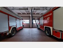 Innenansicht einer Feuerwehrgarage mit zwei roten Löschfahrzeugen, die sich links und rechts befinden. Im Hintergrund sind große Rolltore und eine Abgasabsauganlage sichtbar.