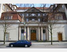 Fassade des Amtsgerichts Tempelhof-Kreuzberg, ein historisches Gebäude mit Säulenportal, symmetrisch angeordneten Fenstern und roten Ziegeldächern; vor dem Gebäude stehen Bäume und ein geparktes Auto.