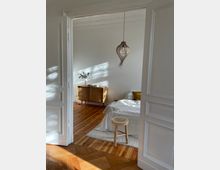 Blick in ein helles Schlafzimmer mit Holzfußboden und weiß gestrichenen Wänden. Im Raum stehen ein Bett mit weißer Bettwäsche, ein kleiner Holz-Hocker, ein Sideboard aus Holz mit Flechtmuster und eine hängende Korblampe; natürliches Licht fällt durch ein Fenster ein.