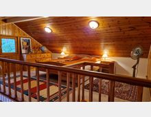 Dachzimmer mit Holzverkleidung an Wänden und Decke, eingerichtet mit einem Doppelbett, Nachttischen, einer Kommode und Teppichen. Das Zimmer hat ein Fenster mit Blick nach draußen, warme Beleuchtung und einen Ventilator in der Ecke.