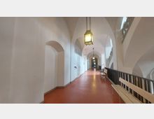 Ein langer, heller Flur mit Gewölbedecke und weißen Wänden, ausgestattet mit hängenden Laternenleuchten. Auf der rechten Seite befindet sich ein schwarzes Geländer sowie helle Holzbänke, die den Flur flankieren, während links Türen und Bögen sichtbar sind.