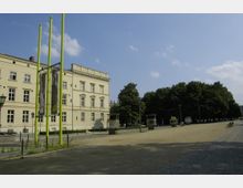 Ein historisches Gebäude mit heller Fassade und mehreren rechteckigen Fenstern befindet sich links im Bild. Davor stehen grüne Fahnenmasten, eine offene, gepflasterte Fläche und rechts ist ein Baumhain zu sehen.