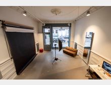 Ein heller Fotostudio-Raum mit schwarzem Hintergrundrollo an der linken Wand, großen Fenstern zur Straße im Hintergrund, einem braunen Sofa auf der rechten Seite und einem Arbeitstisch mit Computer unten rechts. Ein Stativ mit Lichtschirm steht zentral im Raum.